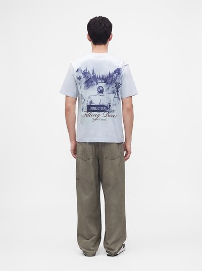 Ένα άτομο που φοράει ένα λευκό T-shirt με σχέδιο θεματικό σκηνοθέτη και χαλαρά καφέ παντελόνια, στεκόμενο με την πλάτη προς τα έξω σε λευκό φόντο.