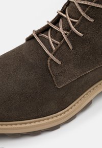 Botín de ante marrón con parte superior texturada, cordones beige y suela de goma. Presenta una puntera redondeada y detalles de costura visibles.