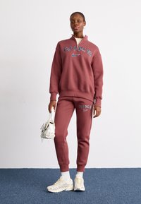 Sweatshirt Nike bordeaux com gola alta e fecho, combinado com calças de treino a condizer. Ténis Nike brancos com detalhes claros, segurando uma mala.
