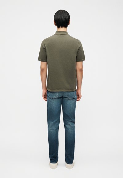 Άνδρας γυρισμένος με την πλάτη, φοράει ελαφρώς πράσινο κοντομάνικο πόλο μπλουζάκι, ξεβαμμένα μπλε τζιν και λευκά αθλητικά παπούτσια πάνω σε μια απλή λευκή φόντο.