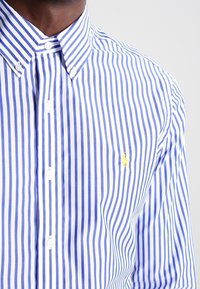Striped button-down shirt featuring thin blue and white stripes, collared neckline, and yellow embroidered logo on the chest. Textured fabric.