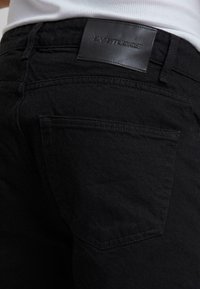 Schwarze Denim-Jeans mit einer glatten Textur, standardmäßigen Gesäßtaschen und einem Lederlabel mit "BY STUDIOS" an der Taille.
