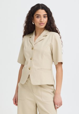 Kvinde med langt krøllet hår iført en beige kortærmet blazer og matchende bukser, stående mod en lys baggrund.