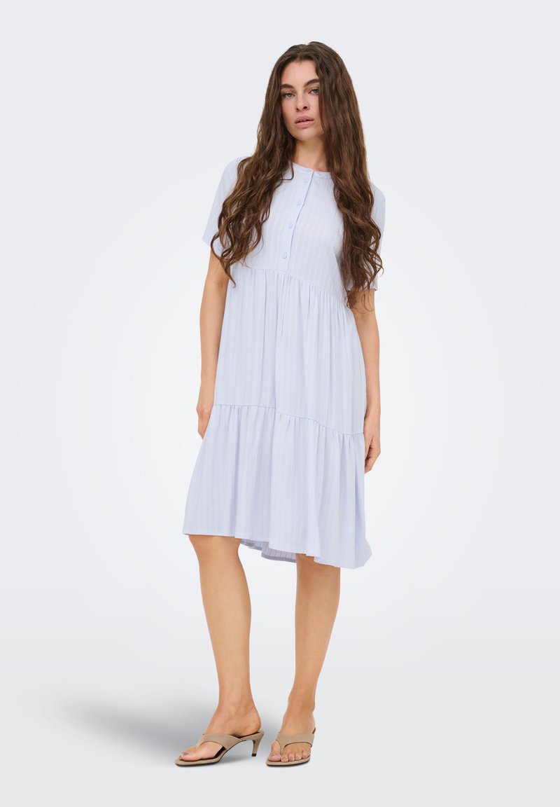 Femme aux longs cheveux bruns portant une robe midi bleu clair à manches courtes et des sandales beiges à talons, debout avec une expression neutre.