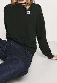 Černá mikina vyrobená z bavlny, s kulatým výstřihem a dlouhými rukávy. Na hrudi je umístěná malá bílá nášivka s logem Lacoste.