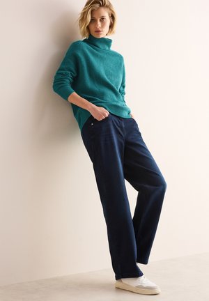 Teal gebreide coltrui met een relaxte pasvorm, gecombineerd met navy blauwe losse broek en witte sneakers. Minimalistische, casual outfit.