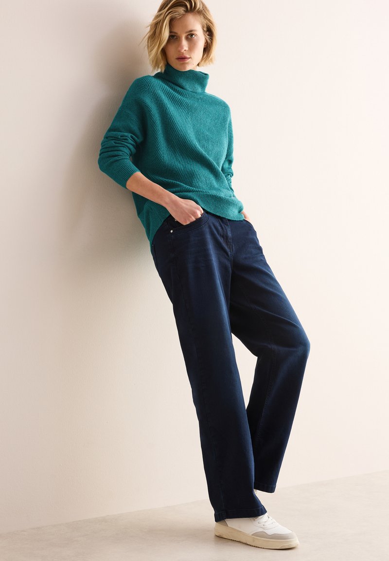 Maglione a collo alto in maglia color turchese con una vestibilità rilassata, abbinato a pantaloni larghi blu navy e sneakers bianche. Outfit minimalista e informale.
