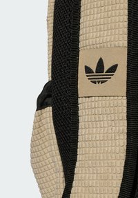Beige textúrájú táska fekete kiegészítőkkel, kiemelkedő felületi mintázattal és egy barna címkével, amelyen az Adidas logo látható. Téglalap alakú.