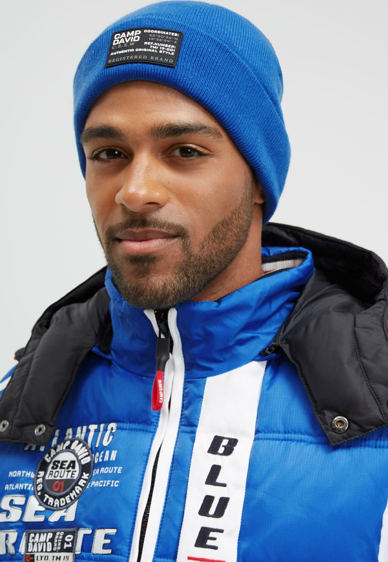Blue insulated jacket with black accents, featuring white stripes and branded patches. The model is wearing a matching blue knit beanie.