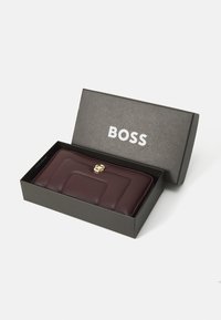 Burgundijas ādas maciņš ar izšūtu dizainu un zelta aksesuāriem, iesaiņots melnā kastē ar baltu BOSS logotipu.