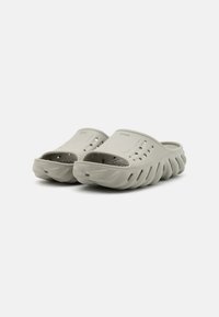Light gray clogs with ventilation holes, contoured footbed, and a thick, wavy sole design. Made from flexible synthetic material.