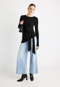 Bluza neagră cu mâneci lungi și detaliu de legare în față, combinată cu blugi evazați bleu deschis și cizme negre cu vârf ascuțit. Geantă de mână neagră ca accent.