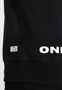 Sudadera negra de algodón con texto "ONE" impreso en blanco en el dobladillo y una pequeña etiqueta de tela con detalles de la marca. Puños acanalados texturizados.