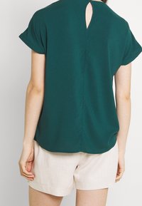 Haut court à manches courtes couleur bleu sarcelle avec une découpe en trou de serrure dans le dos, texture lisse et coupe décontractée, associé à un short en lin de couleur claire.