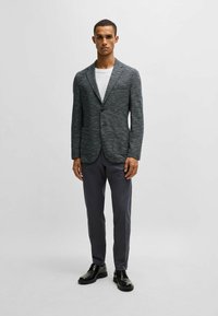 Blazer grigio con texture, colletto a becco, chiusura con un bottone e due tasche frontali, abbinato a pantaloni grigi a vestibilità slim e scarpe nere.