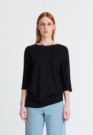 Frau mit glattem roten Haar, die ein schwarzes Langarm-Top, hellblaue Hose, silberne Halskette und Armbänder trägt und vor einem schlichten Hintergrund steht.