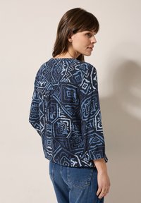 Marinblå topp med ett geometriskt mönster i nyanser av blått, långa ärmar och en avslappnad passform. Tyget är lätt och har en mjuk textur.