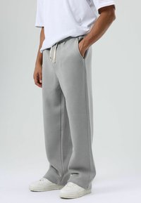 Pantalon de survêtement gris à coupe décontractée, doté de poches latérales et d'une taille élastique avec un cordon beige. Associé à des baskets blanches.