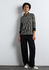 Czarny i kremowy wzorzysty top z wysokim kołnierzem, luźny krój i długie rękawy, w połączeniu z luźnymi czarnymi spodniami i białymi trampkami.