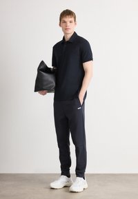 Joven con camiseta de polo negra y pantalones jogger, sosteniendo un gran bolso de cuero negro, vistiendo zapatillas blancas, de pie contra una pared blanca lisa.