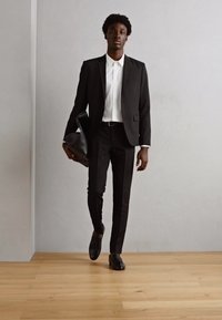 Costum negru bine croit cu o cămașă albă, pantaloni slim-fit și mocasini din piele neagră. Ținând o geantă din piele neagră, stă pe un podea din lemn.