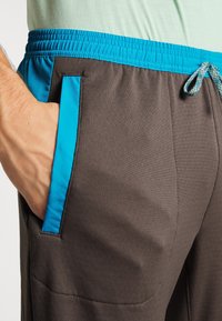 Pantalon de sport gris avec une ceinture turquoise, doté de poches et d'un cordon de serrage. Confectionné en tissu lisse et léger.