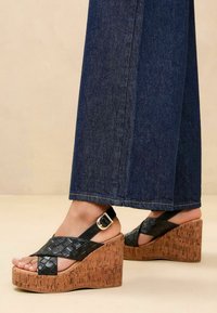 Sandali con tacco a zeppa in pelle intrecciata nera con piattaforma in sughero, caratterizzati da un design a strisce incrociate e chiusura con fibbia regolabile.