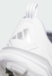 Zapatilla deportiva blanca con parte superior textil lisa, suela de goma y un logo plateado prominente. Incluye un patrón texturizado en la tela.