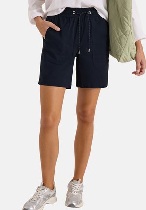 Vrouw draagt zwarte shorts met trekkoord en opgestikte zakken, witte sneakers, wit topje, en houdt een groene doorgestikte jas vast.
