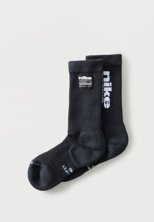 Para czarnych skarpetek Nike typu crew z białym napisem logo i metką na prążkowanych ściągaczach, ułożona płasko na jasnym tle.