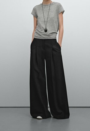 Personne portant une chemise grise à manches courtes, un pantalon noir à jambes larges avec des poches, des chaussures blanches et un long collier pendentif noir.