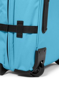 Maleta rodante azul clara con un exterior de tela duradera, ruedas de plástico negro y una correa negra segura. Tiene esquinas reforzadas.