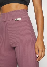 Leggings da allenamento a coste in una tonalità di mauve tenue con una cintura ampia. Presentano un piccolo tag bianco con il logo Adidas sulla cintura. Texture morbida.