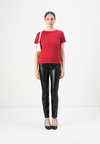Woman standing, wearing red short-sleeve top, black leather pants, black heels, and carrying a small white shoulder bag.