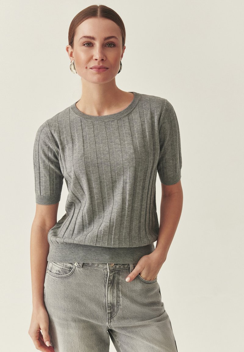 Pull gris en maille côtelée avec manches courtes et taille ajustée, associé à un jean gris clair. Texture lisse avec de fines lignes verticales.