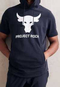 Laivastonsininen hupullinen lyhythihainen collegepaita, jossa on valkoinen härkälogo ja teksti "PROJECT ROCK". Valmistettu pehmeästä, hengittävästä kankaasta, jossa on rento istuvuus.