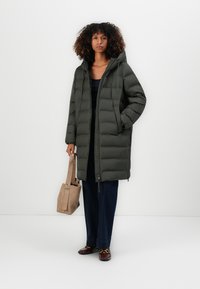 Dunjakke i mørkegrønn med glidelås foran og hette, med quiltet design. Modellen holder en beige tote bag, og har på seg mørke bukser og topp.