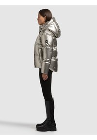 Donna con giacca imbottita argento con cappuccio, pantaloni neri e stivali neri sopra il ginocchio, in piedi di profilo su uno sfondo semplice.