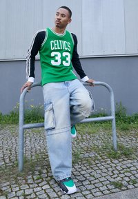 Grünes Netz-Basketballtrikot mit "CELTICS" und der Nummer "33", kombiniert mit übergroßen hellblauen Cargo-Jeans und grünen Sneakers.