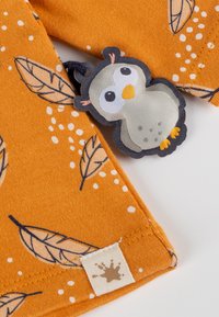 Orangener Baumwollstoff mit Blattmustern und weißen Punkten, mit einem grauen Stoff-Label, das mit einem dunkelblauen Faden befestigt ist.