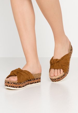 Des pieds portant des sandales à plateforme en suédé marron avec des lanières nouées et des détails en imprimé léopard sur les semelles épaisses.