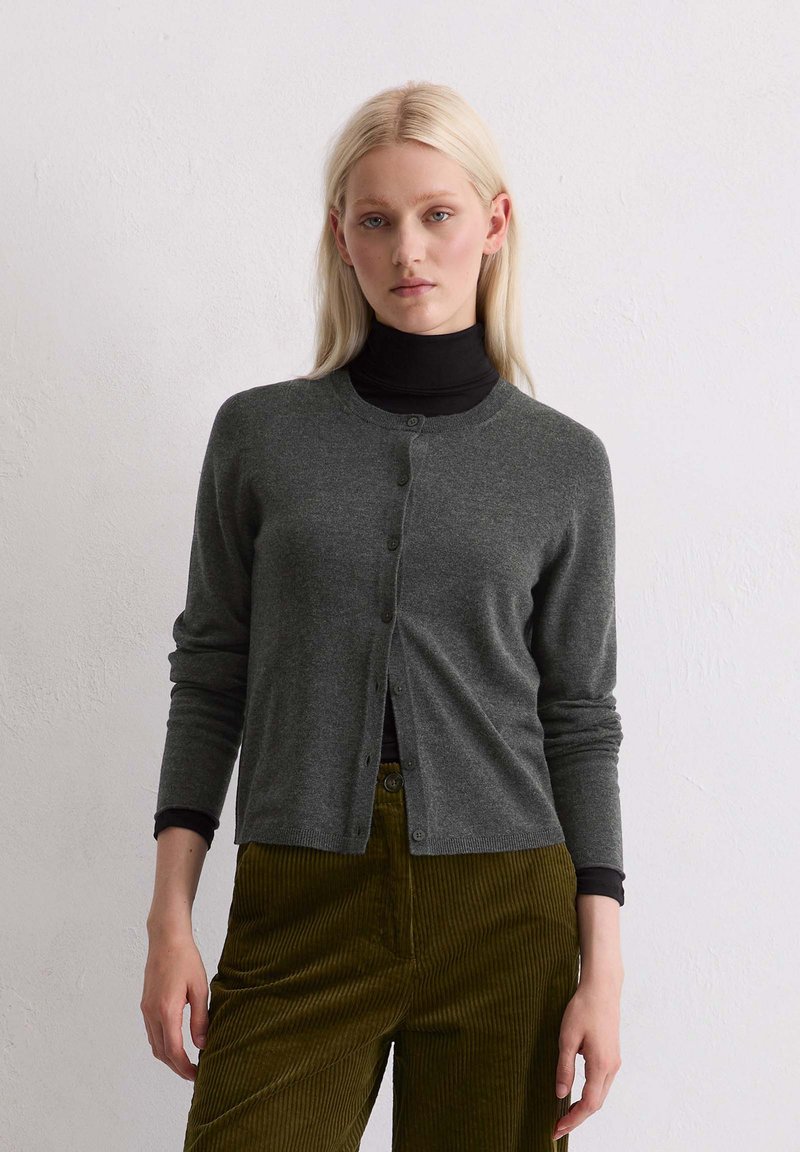 Cardigan gris en matière fine tricotée avec un col rond, des boutonnières à l'avant et des manches longues, porté sur un col roulé noir et un pantalon en velours côtelé.