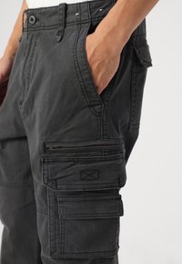 Detailní záběr na černé cargo kalhoty s několika kapsami, včetně kapsy na zip, které má na sobě osoba s rukou v boční kapse.