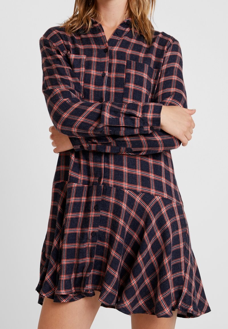 Femme portant une robe boutonnée à manches longues à carreaux bleu marine et rouge avec une jupe évasée, les bras croisés sur la poitrine.