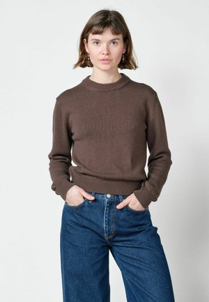 Femme aux cheveux courts et bruns portant un pull marron et un jean bleu, debout les mains dans les poches devant un fond clair uni.
