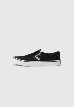 Sapatilha preta de lona slip-on com costura branca, sola de borracha branca e uma pequena etiqueta de logotipo. Textura suave, design minimalista.