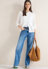 Femme debout contre un mur beige, portant une chemise blanche à boutons, un jean bleu, des baskets, tenant un grand sac à main en daim marron dans la main droite.