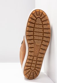 Semelle de chaussure marron avec des crampons profonds et le logo ECCO, inclinée contre des murs et un sol blancs.