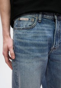 Ανοιχτό μπλε τζιν με ξεθωριασμένο φινίρισμα και αντίθετες ραφές. Μια κόκκινη και λευκή ετικέτα «Calvin Klein» είναι ορατή κοντά στη μέση.