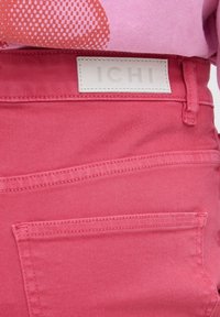 Prim-plan cu buzunarul din spate și talia blugilor roz din denim, cu etichetă albă de brand ICHI, asortați cu o cămașă roz cu model.